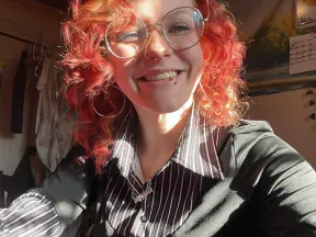 A selfie of a person with curly red hair and glasses smiling into the camera.