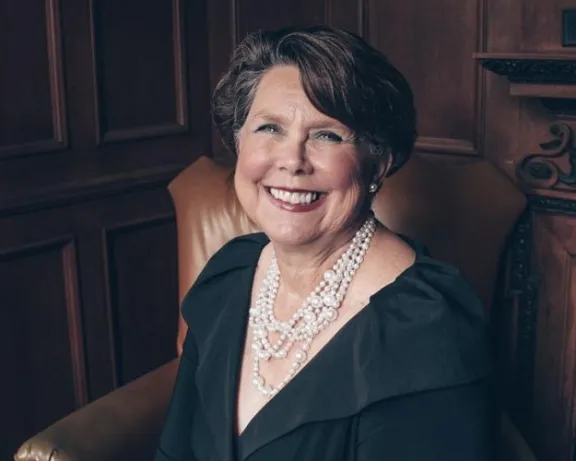 Smiling woman in black dress and pearls seated in leather armchair, wood-paneled room.